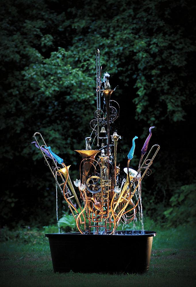 Musical instrument fountain sculpture constructed from copper tubing, a water wheel, blown glass and a group of upcycled, brass musical instruments including saxophones, trombones, trumpets, and a tuba.